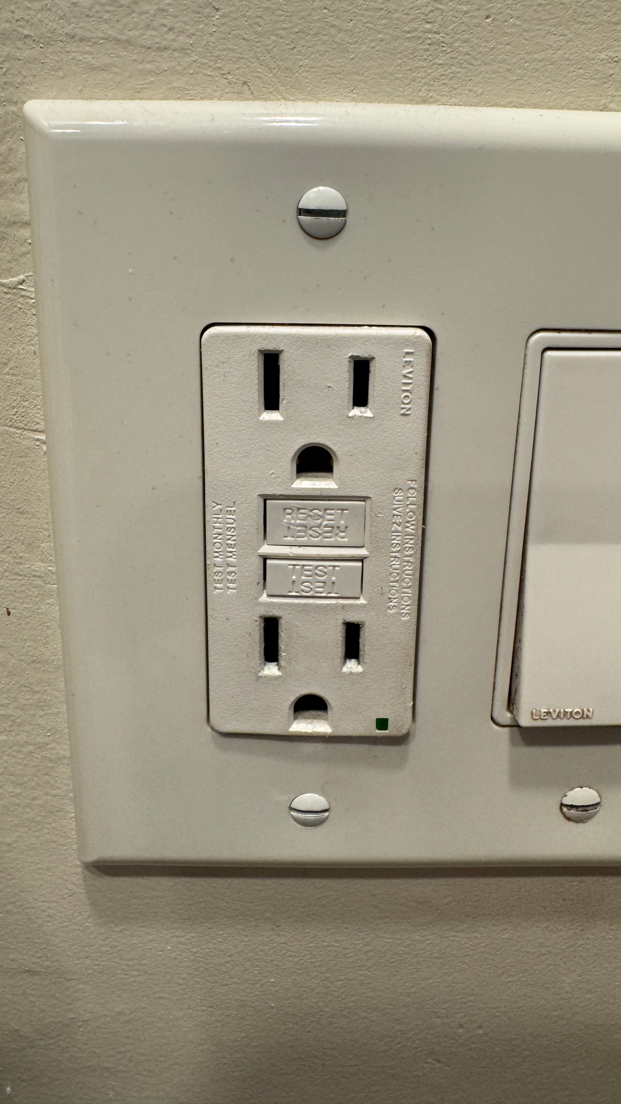 Close-up photograph of a white Leviton GFCI receptacle showing the TEST and RESET buttons and green indicator light
